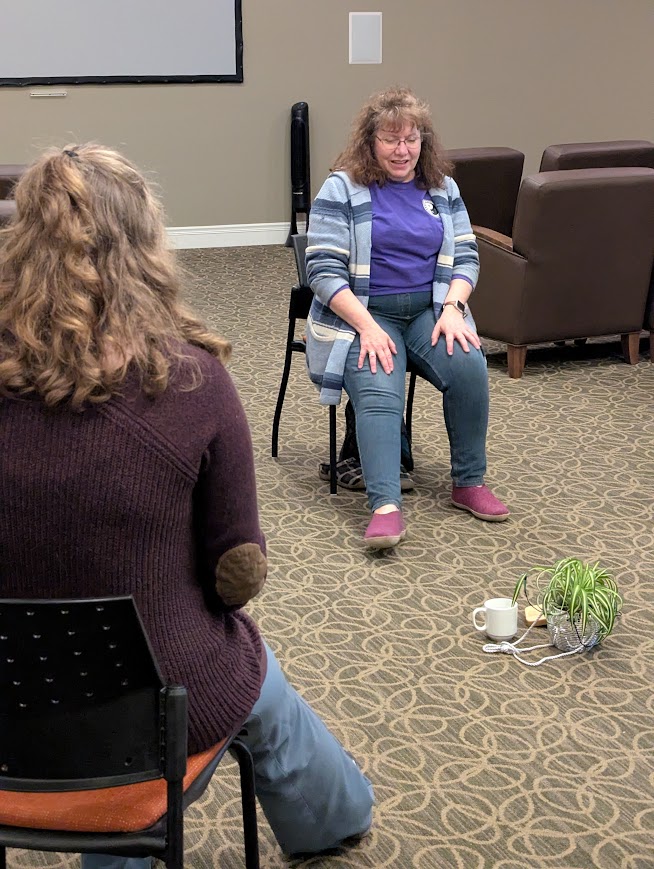 woman sitting on a chair and turning foot while a student copies across the room, a plant is on the floor between them